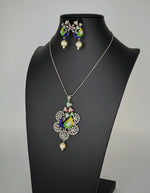 Silver polished a peacock pendant with vibrant green, blue, and gold enamel accents, along with sparkling embellishments, accompanied by matching earrings, displayed on a black mannequin necklace stand.