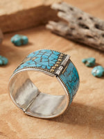 A wide, hinged cuff bracelet made of sterling silver, featuring an intricate mosaic pattern of fragmented blue turquoise stones and decorative gold-tone studs, standing upright on a textured sandstone surface with blurred Southwestern-themed items in the background.