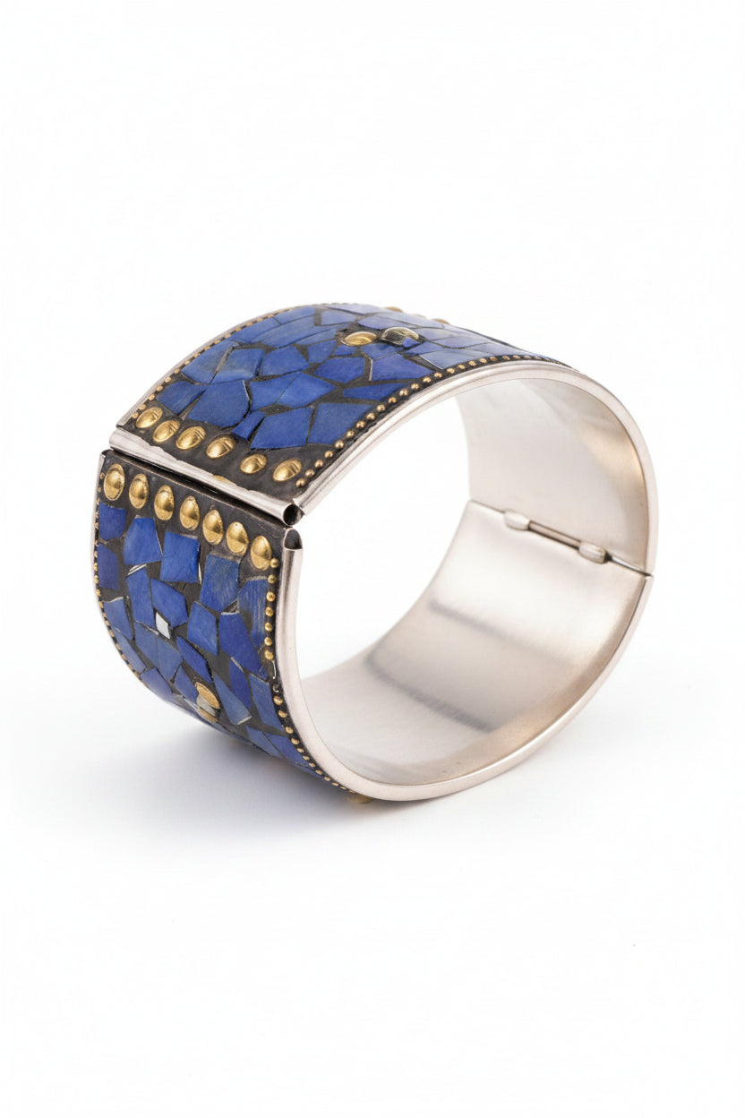A wide silver-toned cuff bracelet with a textured blue mosaic inlay and rows of small gold studs, photographed on a dark fabric background.