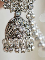 Silver-toned ornate jhumka earrings with clear rhinestones and dangling bead chains, shown in a close-up studio shot.
