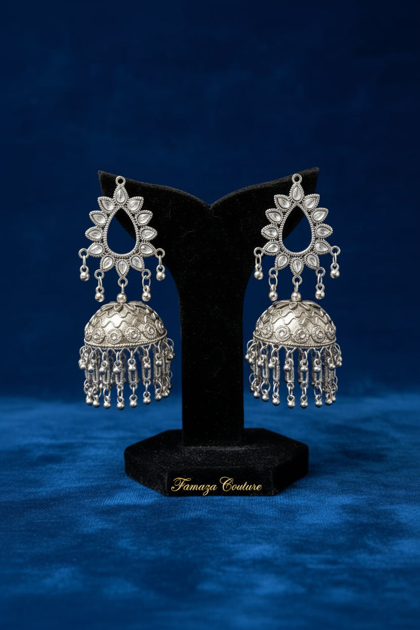 A pair of ornate silver-toned jhumka earrings displayed on a black velvet stand, set against a softly blurred warm-toned background.