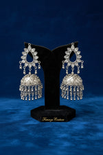 A pair of ornate silver-toned jhumka earrings displayed on a black velvet stand, set against a softly blurred warm-toned background.