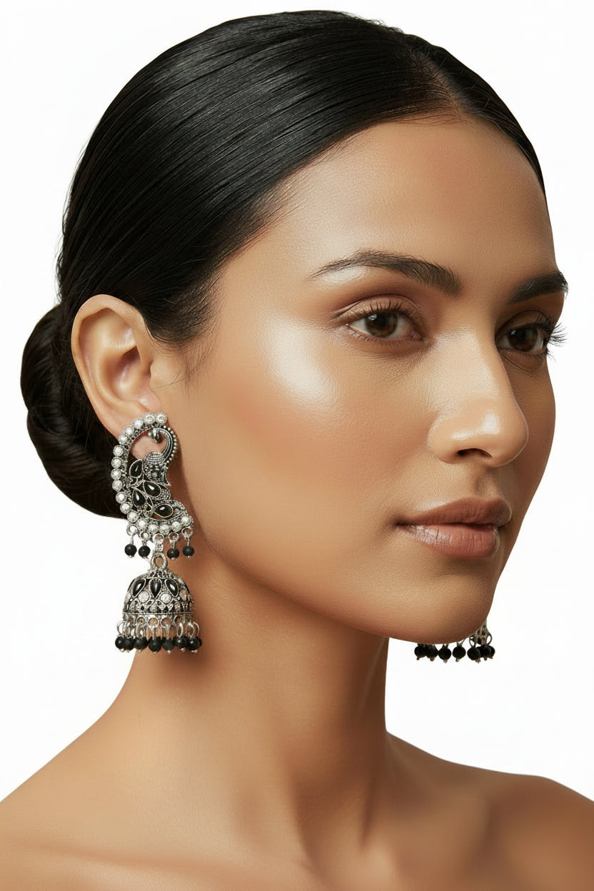 A woman with her hair in a low bun models a pair of ornate silver-toned Indian jhumka earrings with black and pink stone accents, set against a classic cream-colored wallpaper background.