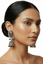 A woman with her hair in a low bun models a pair of ornate silver-toned Indian jhumka earrings with black and pink stone accents, set against a classic cream-colored wallpaper background.
