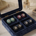 A navy blue velvet jewelry box with a glass lid, displaying six pairs of stud earrings in different colors and metal settings, placed on a light marble surface.