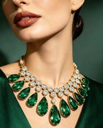 Close-up view of Emerald Cascade Teardrop Statement Necklace featuring cascading pear-shaped emerald green gemstones surrounded by brilliant round clear crystals, set in polished gold-tone finish, worn on neck against deep green background.