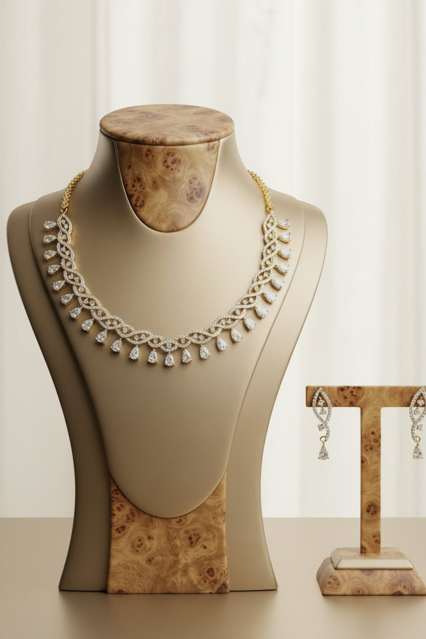 A gold and diamond necklace displayed on a beige jewelry bust, with matching dangling earrings on a stand beside it against a dark marble background.