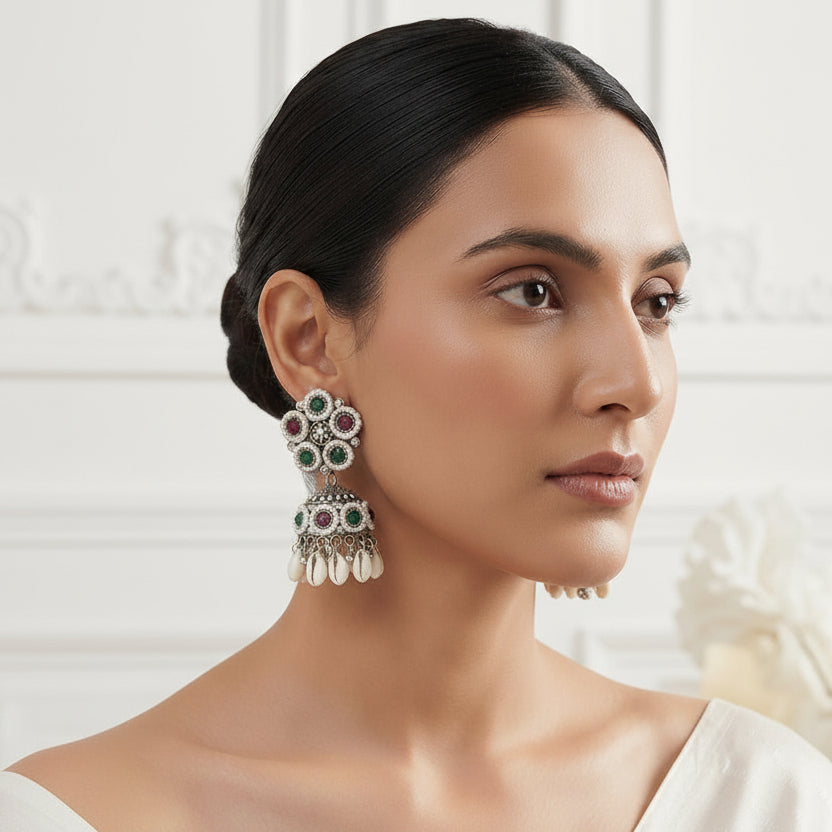 A woman wearing ornate silver-toned Indian jhumka earrings with green, red, and white stones, shown in a close-up portrait against a soft white background.