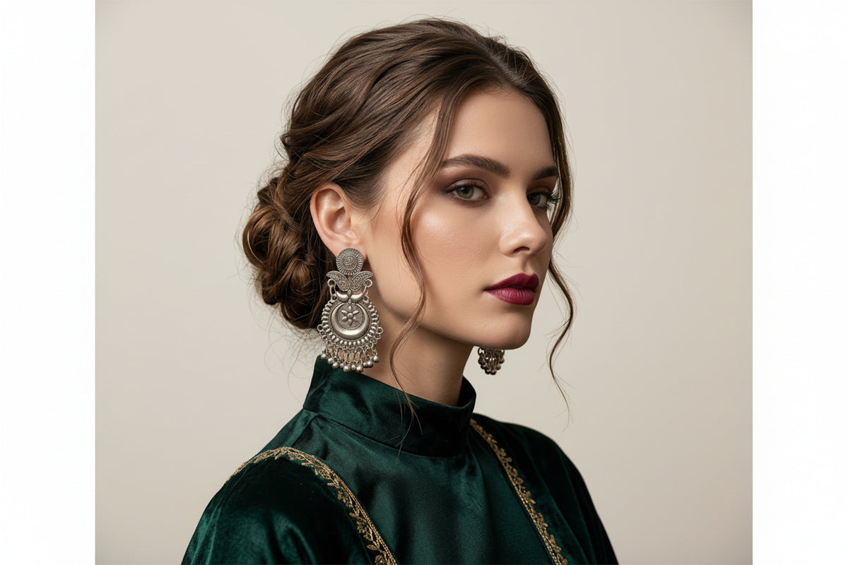 A woman with styled brown hair in an updo wearing a dark green velvet high-neck top with gold embroidery, showcasing a large, ornate silver-toned traditional Indian jhumka earring.
