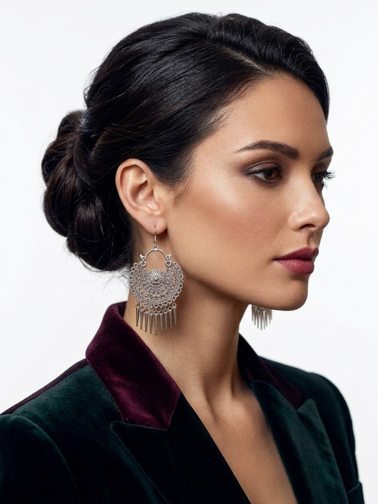 A woman in profile wearing large silver-toned filigree dangle earrings with fringe, paired with a dark green velvet jacket featuring a burgundy collar, against a plain light gray background.
