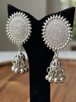 A pair of intricate silver-toned Indian jhumka earrings displayed on a black velvet stand. The earrings feature circular, heavily textured mandala designs with dangling bell-shaped charms.