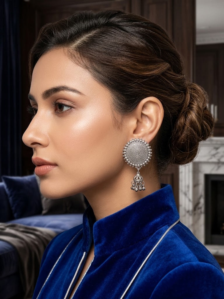 A woman in profile wearing a large silver-toned jhumka earring with a textured circular face and dangling charm, paired with a royal blue velvet jacket in an indoor setting.
