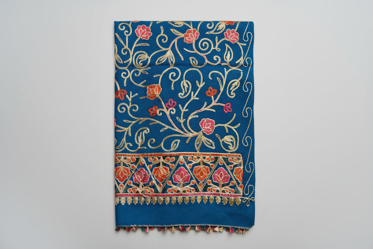 The blue Kashmiri embroidered pashmina scarf with tassels hanging at the ends, displayed on a wooden surface.
