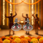 The pair of silver toned metal oxidised earrings design is showing a woman is playing dandiya in navratri lehnga choli traditional dress in red,white and black colour for navratri nights.
