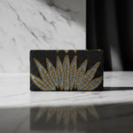 The rear view of this handcrafted clutch bag with a geometric pattern of glass beads in gold and blue on a black base, displayed on a wooden surface with green leaf accents around.