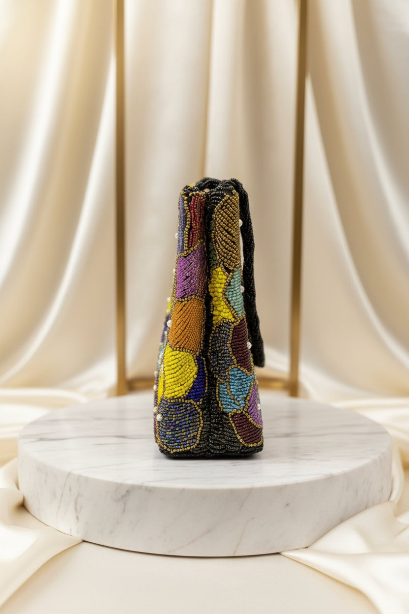 The side view of A multi-coloured circular handcrafted tote bag with pearl embellishments, displayed against a leafy backdrop.