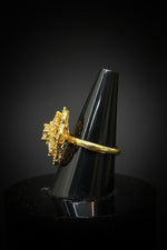 The side view of a gold ring with a large, ornate baguette crystal cluster setting, displayed on a black cone ring stand held by a hand.