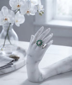 The silver ring with a green and white floral cluster setting, displayed on a white marble hand-shaped mannequin. The scene is set on a white marble table with white orchids in a glass vase in the background.