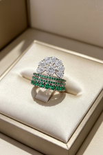 A silver-tone ring set with a large clear floral cluster top and two rows of green gemstones, displayed on a cream satin ring box.