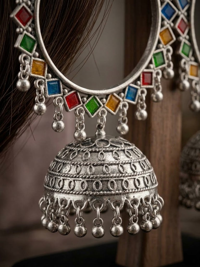A close-up of a silver-toned traditional Indian jhumka earring with colorful square gemstones in red, green, blue, and orange, hanging from a silver-toned circular band. The earring is shown worn on a person's ear with dark hair visible, against a warm brown background.