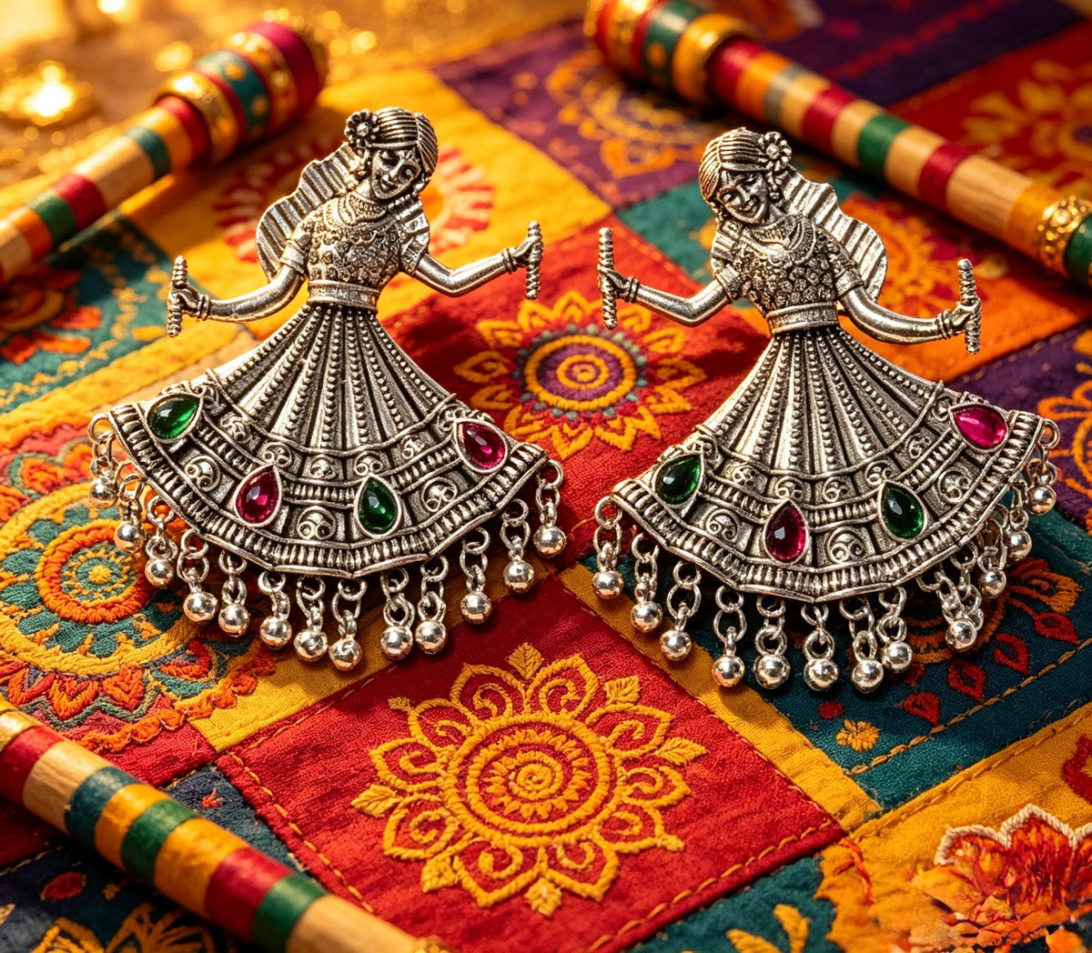 Two ornate silver-toned jhumka earrings shaped like dancing figures, decorated with green and red stones and dangling bells, displayed on a colorful embroidered textile with wooden sticks.
