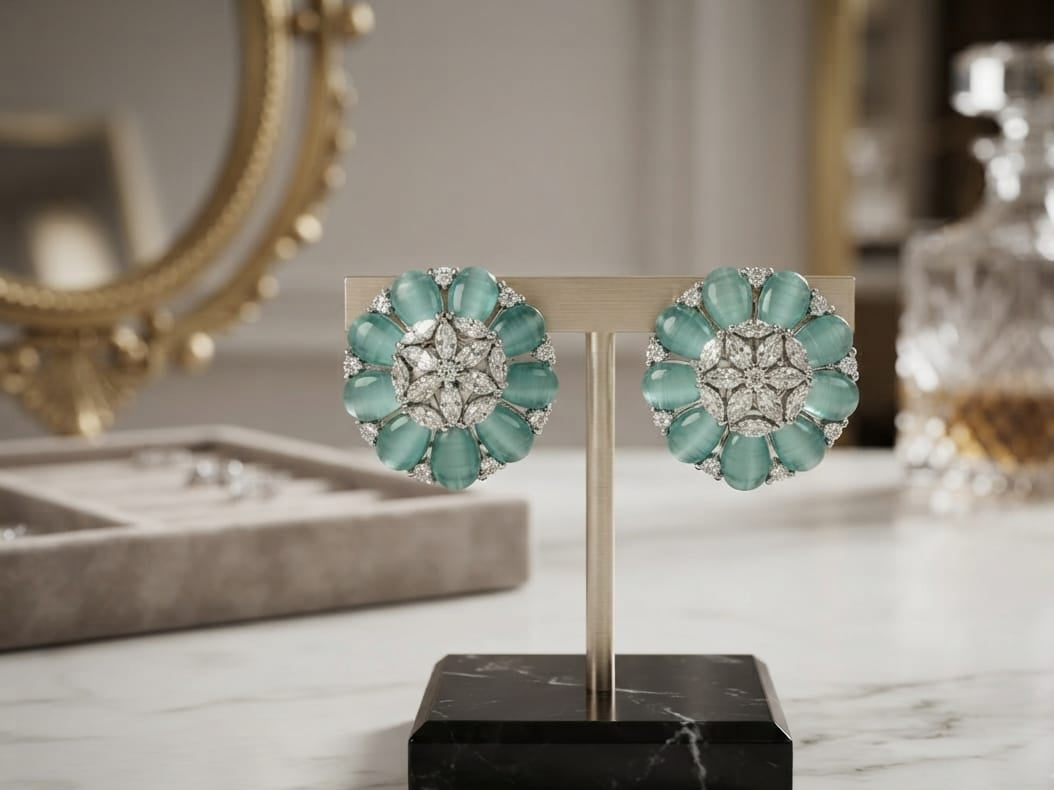 A pair of floral cluster earrings displayed on a stand on a marble surface. The earrings feature mint green marquise-cut stones and clear round stones in a silver-toned setting, with a blurred luxury background including a gold mirror frame and a crystal decanter.