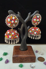 A pair of multicolor beaded Indian jhumka earrings displayed on a dark tree-shaped jewelry stand. The earrings feature intricate silver-toned filigree borders, colorful seed bead patterns in pink, yellow, orange, green, and white, and dangling silver-toned bell charms.