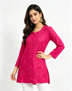 A woman modeling a bright pink embroidered short kurti with white pants standing against a light grey studio background.