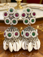 A pair of traditional Indian Jhumka earrings with silver-toned metalwork, white faux pearls, green and magenta stones, and dangling white cowrie shells, displayed on a wooden surface.