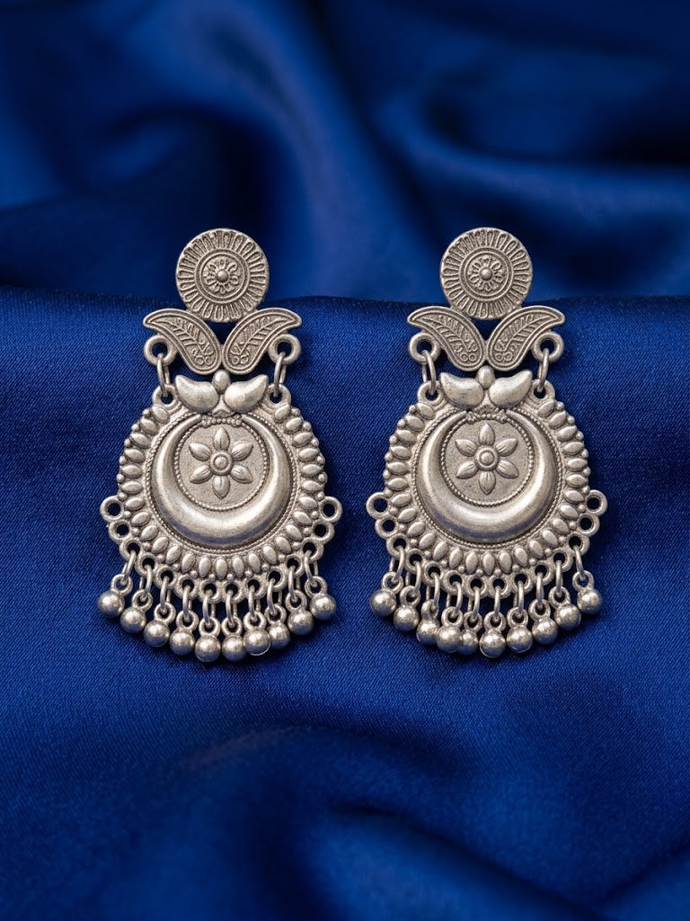 A pair of ornate silver-toned traditional earrings with intricate filigree, floral motifs, and dangling bead chains, displayed on a draped royal blue fabric background.