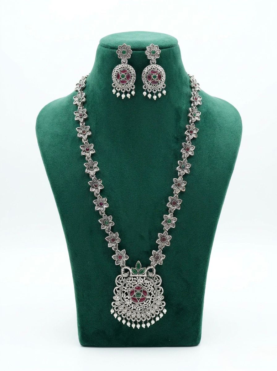 Long oxidized silver necklace and matching earrings with floral patterns and pink and green stones displayed on a green velvet stand.