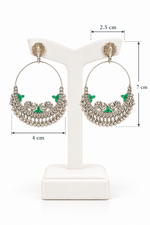A pair of oxidized silver hoop earrings featuring irregular textured studs, traditional peacock motifs with vibrant green stone accents, and hanging bead tassels, displayed on a black stand