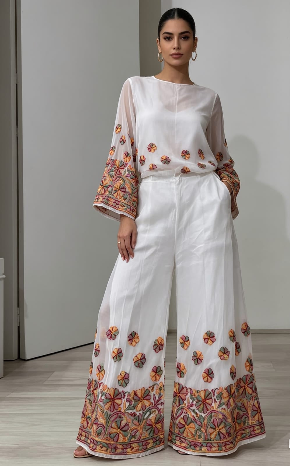 A close-up photograph focusing on the lower half of a woman wearing flowing, white wide-leg palazzo pants. The pants feature intricate, multi-colored floral embroidery along the entire bottom hem and scattered as smaller motifs across the lower leg area. The upper part of her body and the top are cropped out of the frame, emphasizing the detailed design of the palazzo pants as they hang loosely. She is standing on a light wood floor against a plain white background. The lighting is soft and even.