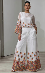 A close-up photograph focusing on the lower half of a woman wearing flowing, white wide-leg palazzo pants. The pants feature intricate, multi-colored floral embroidery along the entire bottom hem and scattered as smaller motifs across the lower leg area. The upper part of her body and the top are cropped out of the frame, emphasizing the detailed design of the palazzo pants as they hang loosely. She is standing on a light wood floor against a plain white background. The lighting is soft and even.