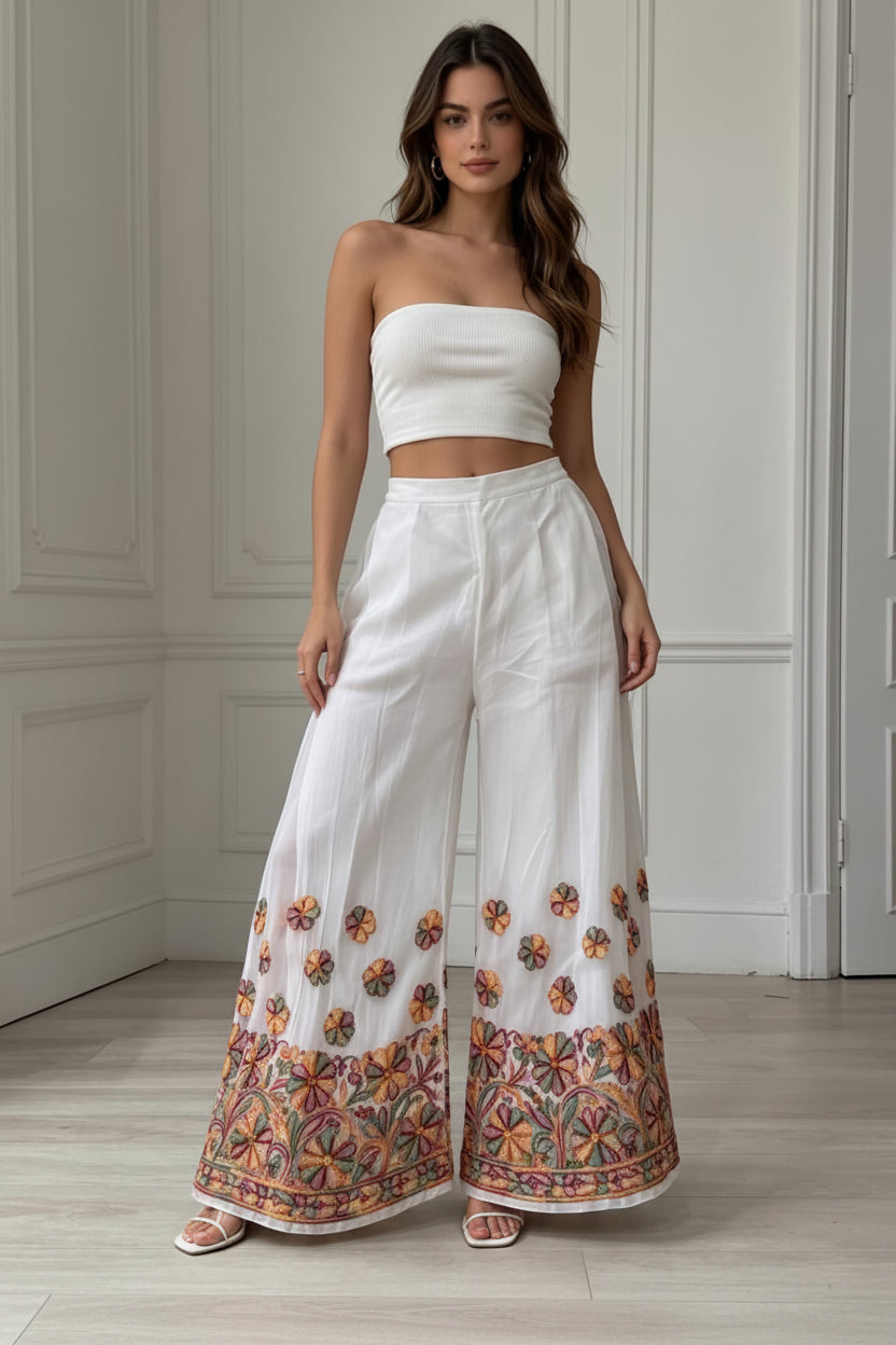 A close-up photograph of the lower half of a woman wearing flowing, wide-leg white palazzo pants. The pants feature detailed, multi-colored floral embroidery across the bottom hem and lower leg section, with smaller floral motifs scattered above the main border. She is standing on a light wood floor, and the upper part of her body and the top she is wearing are cropped out of the frame. The pants are the central focus.