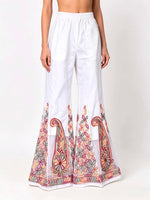 A front view of a model wearing high-waisted white semi-georgette sharara pants with a fluid, wide-leg flared silhouette, featuring a sheer white organza panel at the bottom hem decorated with intricate multi-color traditional paisley and geometric embroidery in rust red, sage green, and pastel yellow.