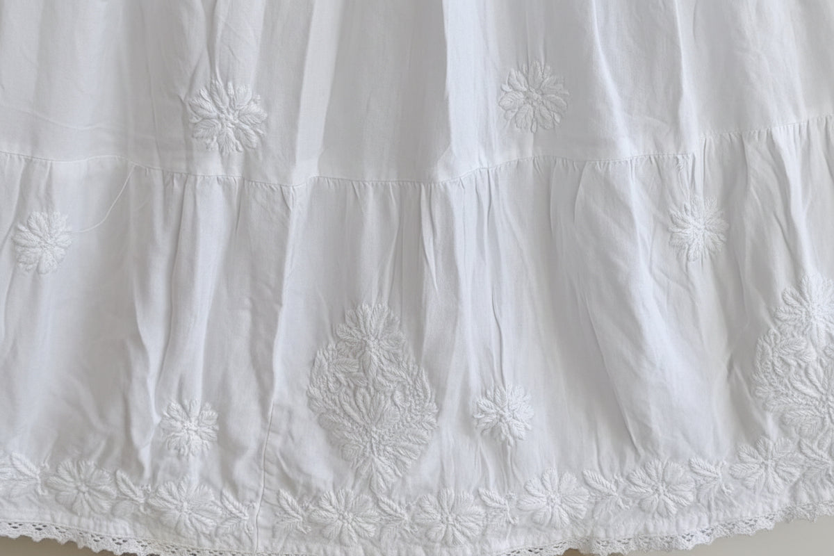 White embroidered cotton fabric with floral and geometric embroidery, featuring a ruffled tiered hem and lace trim.