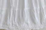 White embroidered cotton fabric with floral and geometric embroidery, featuring a ruffled tiered hem and lace trim.