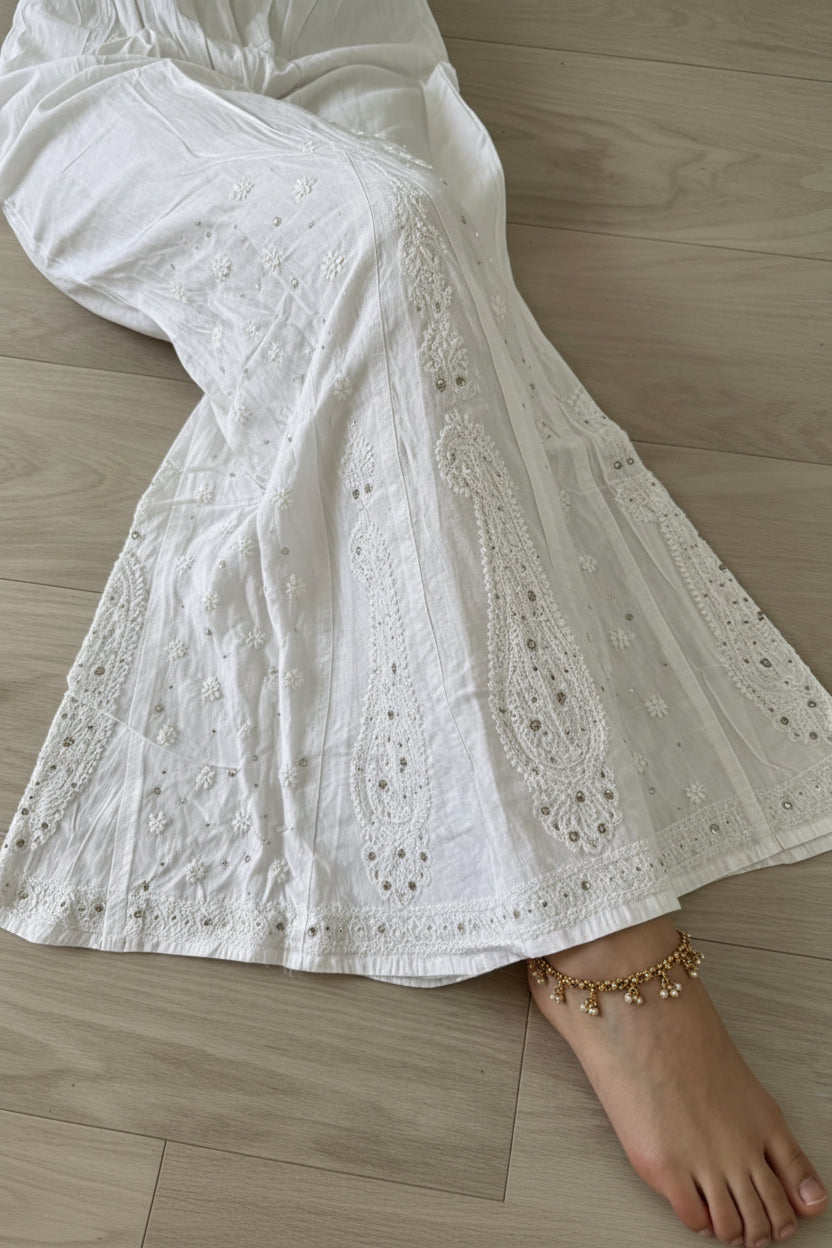 White embroidered cotton flare pants with eyelet detailing, shown on a model lying on a light wood floor.