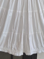 White tiered skirt with delicate floral embroidery and lace trim along the hem, shown hanging against a dark background.