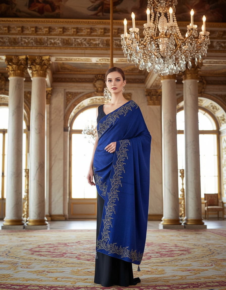 Woman in a blue velvet shalwl wrapping it on with intricate designs standing in an opulent room with chandeliers.