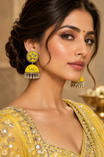 A close-up portrait of a woman wearing ornate yellow and silver jhumka earrings with a yellow embellished traditional outfit against a warm beige background.