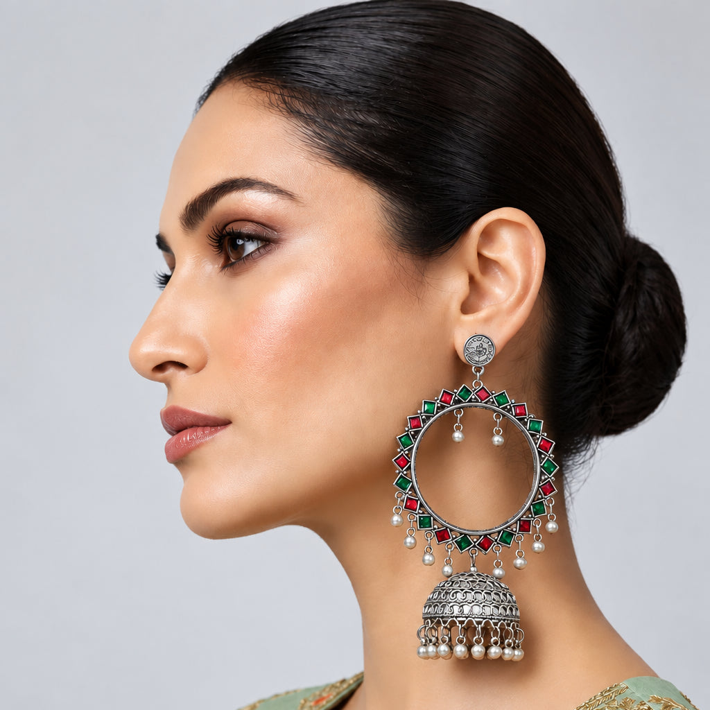 A woman in profile wearing a large silver-toned jhumka earring embellished with green and red enamel paint, paired with a light green embroidered traditional outfit against a neutral gray background.