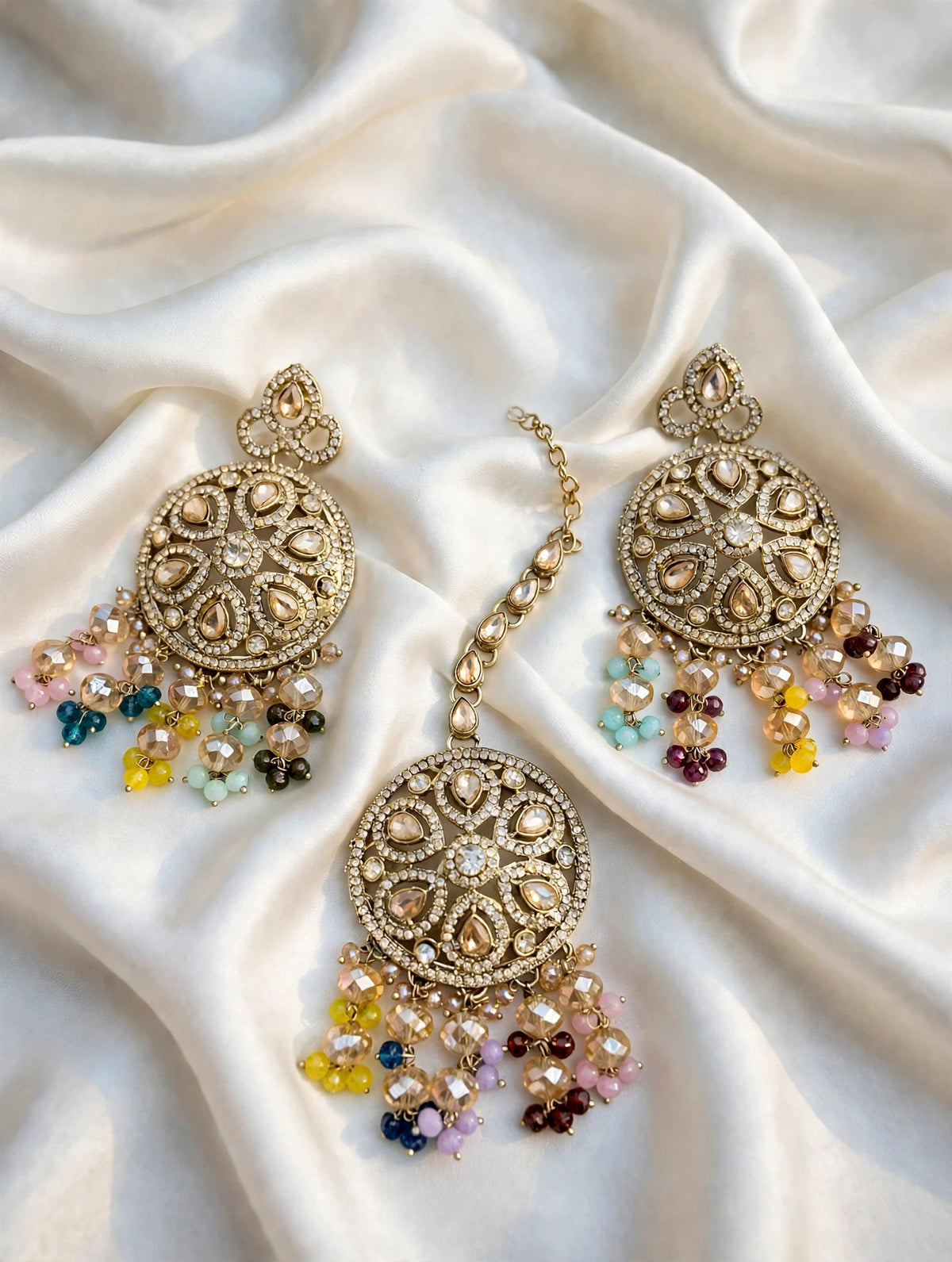 A pair of ornate gold-toned Indian jhumka earrings with intricate beadwork and dangling multicolor beads, displayed on a draped cream satin fabric.