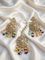 A pair of ornate gold-toned Indian jhumka earrings with intricate beadwork and dangling multicolor beads, displayed on a draped cream satin fabric.