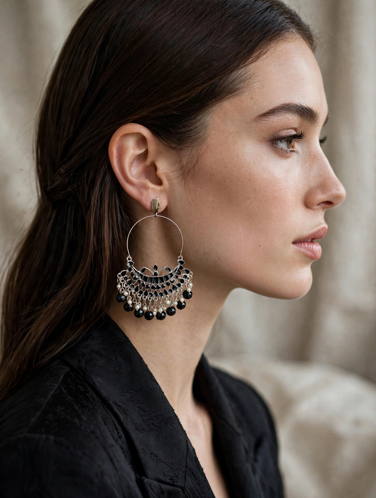 A close-up side profile of a woman wearing a large silver-toned huggie hoop earring adorned with dangling black beads and metallic fringe, set against a soft beige background.