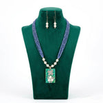 blue beaded necklace with a rectangular pendant and matching earrings on a green velvet stand.