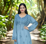 A woman standing in a garden path, wearing a blue Chikankari short kurti frock with floral embroidery.