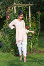 A woman standing outdoors wearing a short frock style kurti with embroidery and a tie-up waist, in a garden showing full look with trousers