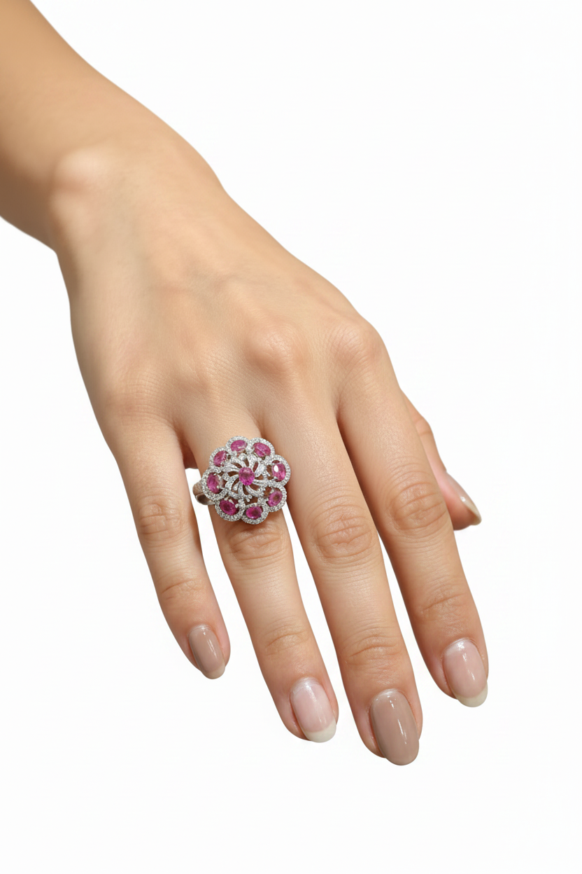 A close-up of a hand wearing a floral cluster ring with pink gemstones and a silver-toned setting, photographed against a plain light gray background.